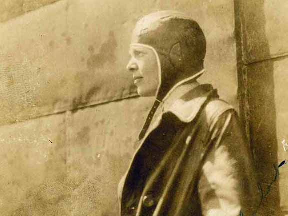 A person wearing an aviator cap and coat stands beside an aircraft. The image has a vintage, sepia-toned appearance.