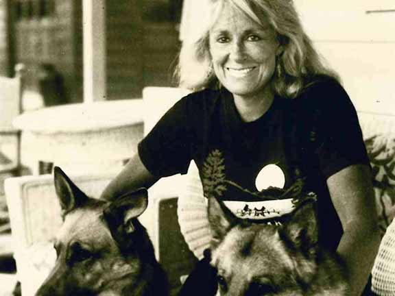 A person smiling while sitting with two German Shepherd dogs on a porch. Casual setting with wooden furniture in the background.