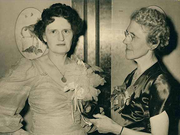 Two people stand indoors; one holds a flower bouquet and a small object. Both wear formal attire. Background shows framed floral artwork.