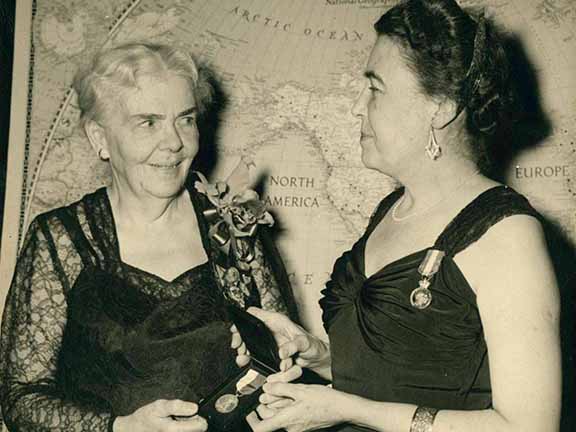 Two persons in formal attire exchange an award in front of a world map. One holds a medal while the other smiles.