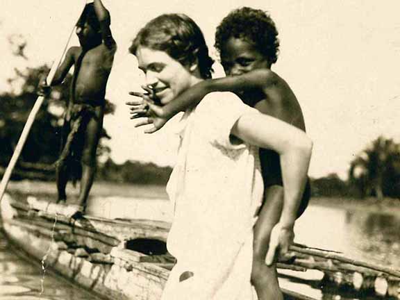 A person smiles with a child on their back in a canoe, guided by another person with a pole on a river.