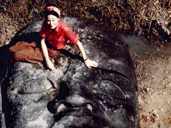 A person in red shirt sits on a large, ancient stone face partially buried in the ground with surrounding grass and soil.