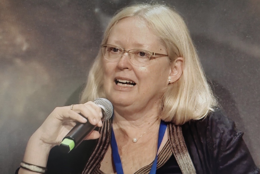 A person with light hair and glasses speaks into a microphone, wearing a blue lanyard. The background is out of focus.