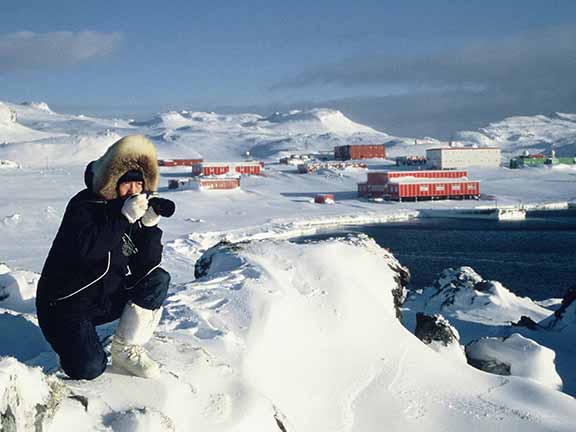 A person in winter clothing photographs snowy Antarctic landscape, with research station buildings visible under a clear sky.