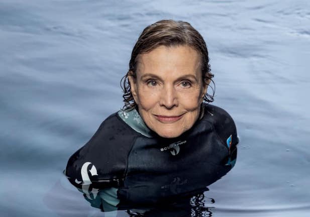 A person in a wetsuit is partially submerged in calm water, looking directly at the camera with a composed expression.