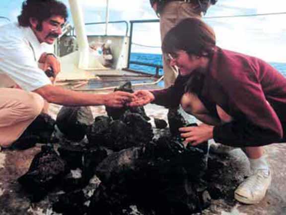 Two people kneel on a ship's deck examining dark rocks, possibly geological samples, with the ocean in the background.