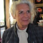 A person with white hair, wearing a plaid jacket, sits indoors near a window. Abstract art is visible in the background.