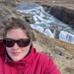 A person in a red coat takes a selfie with Gullfoss waterfall in the background, under a partly cloudy sky.