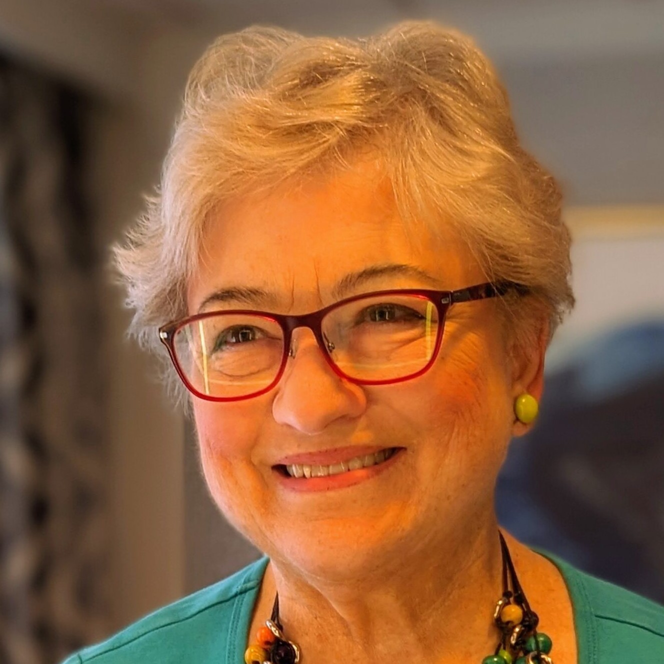 A person with short hair and glasses smiles, wearing a colorful necklace. Bright room with a blurred painting in the background.