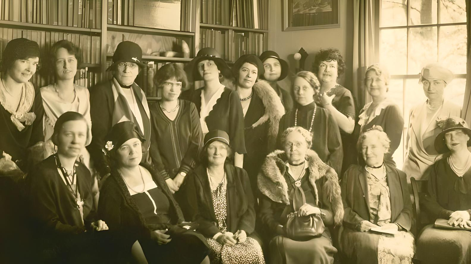A group of people dressed in early 20th-century attire pose indoors, surrounded by bookshelves, conveying a formal, historical gathering.