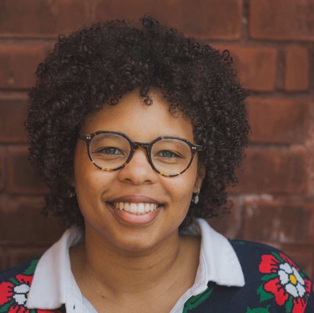 A person with curly hair and glasses smiles, wearing a floral sweater, standing against a brick wall background.