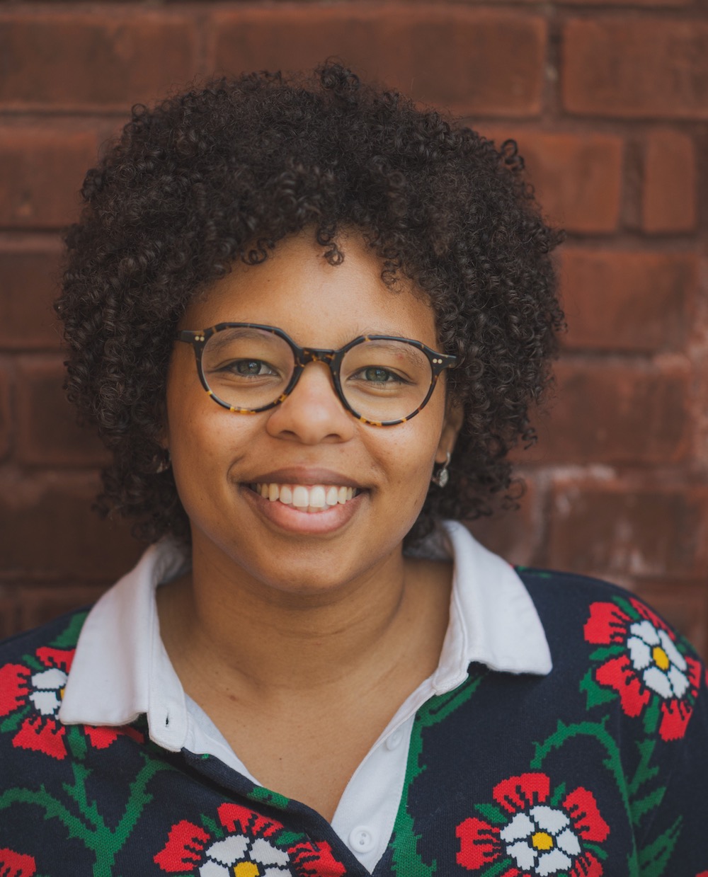A person with curly hair and glasses smiles against a red brick wall, wearing a flower-patterned sweater. No landmarks are visible.