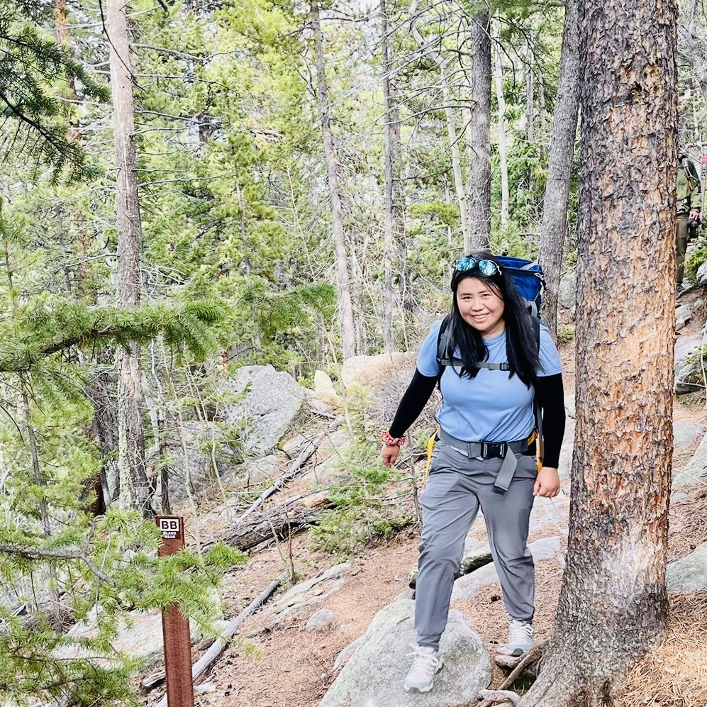 A person hikes through a wooded trail surrounded by dense trees. They carry a blue backpack and smile, enjoying the natural scenery.