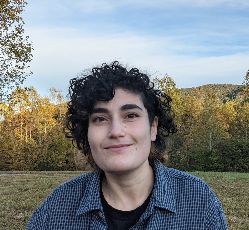 A person with curly hair smiles outdoors, surrounded by autumn trees and mountains under a partly cloudy sky.