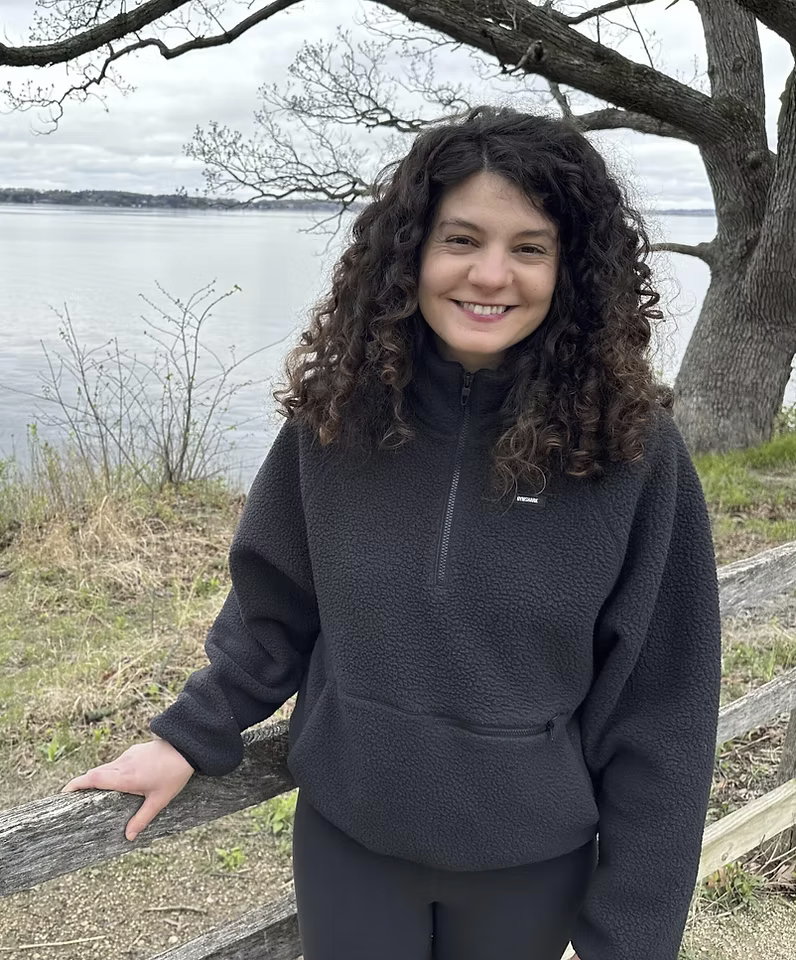 A person with curly hair is smiling beside a wooden fence, with a tree and a large body of water in the background.