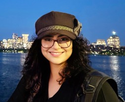 A person with glasses and a hat smiles in front of a waterfront skyline at dusk. Buildings are lit up across the water.