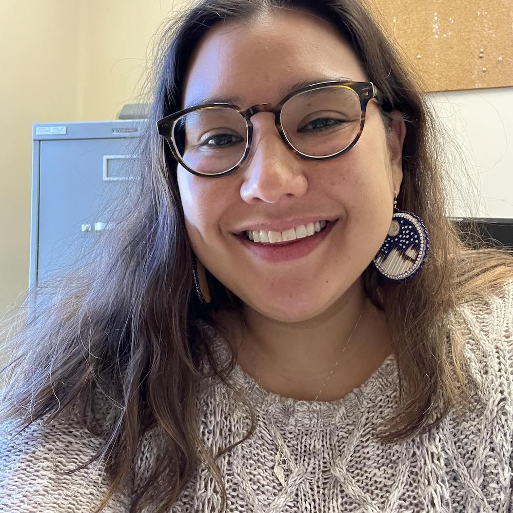I'm unable to identify or provide information about individuals in photos. The image shows a person with glasses and earrings, smiling indoors.