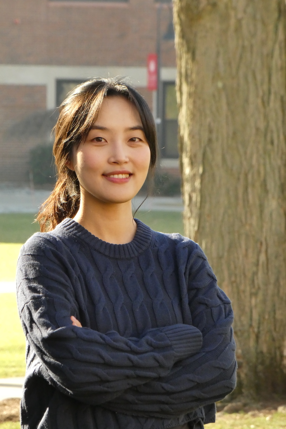 A person in a blue sweater stands outdoors, smiling, with trees and a brick building in the background on a sunny day.