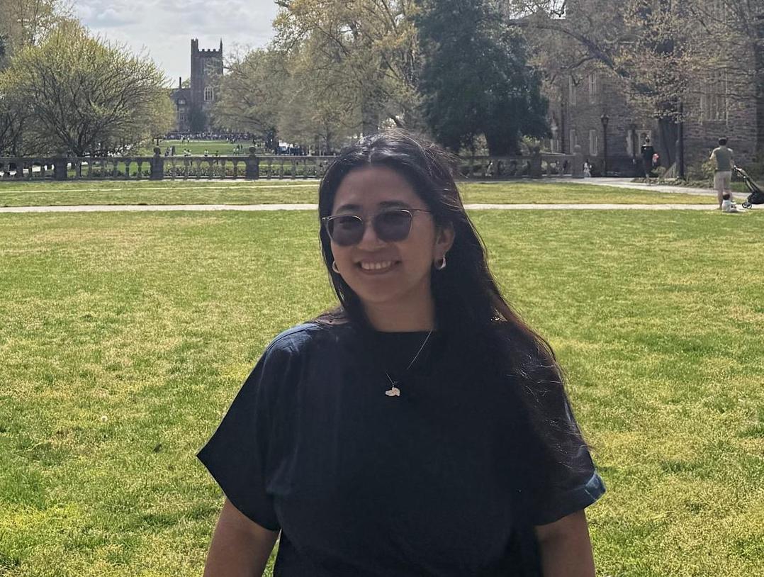 A person stands smiling on a grassy field, with a historic building resembling Princeton University in the background, under a partly cloudy sky.