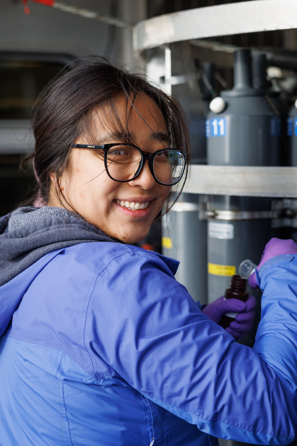 A person in a blue jacket smiles while working with scientific equipment, wearing glasses and gloves, in a laboratory setting.