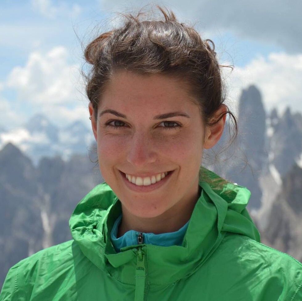 A person in a green jacket smiles, with majestic mountain peaks, possibly the Dolomites, in the background under a cloudy sky.